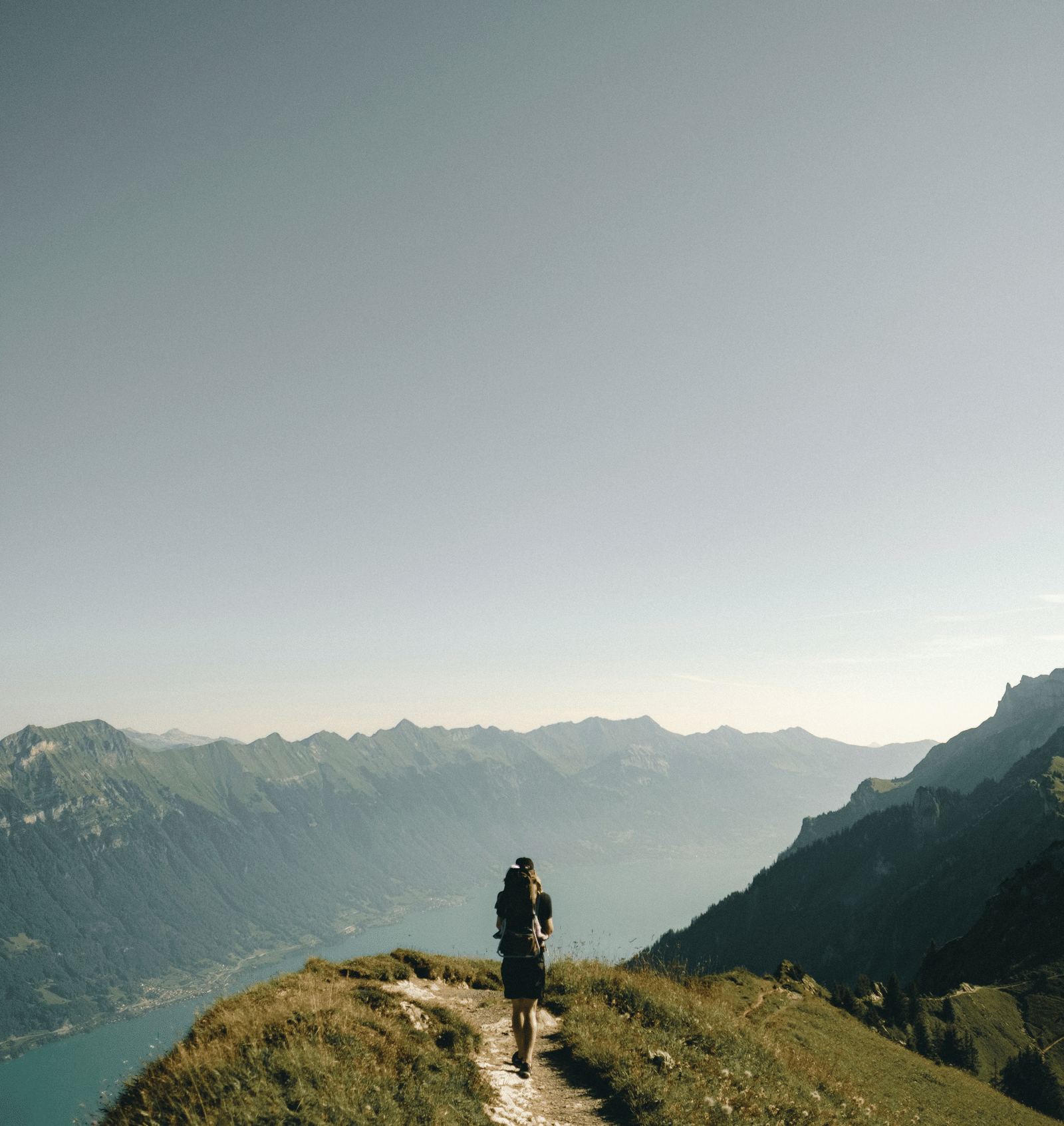 A lone hiker standing on a grassy mountain ridge looking down at a vast blue lake nestled in a valley.