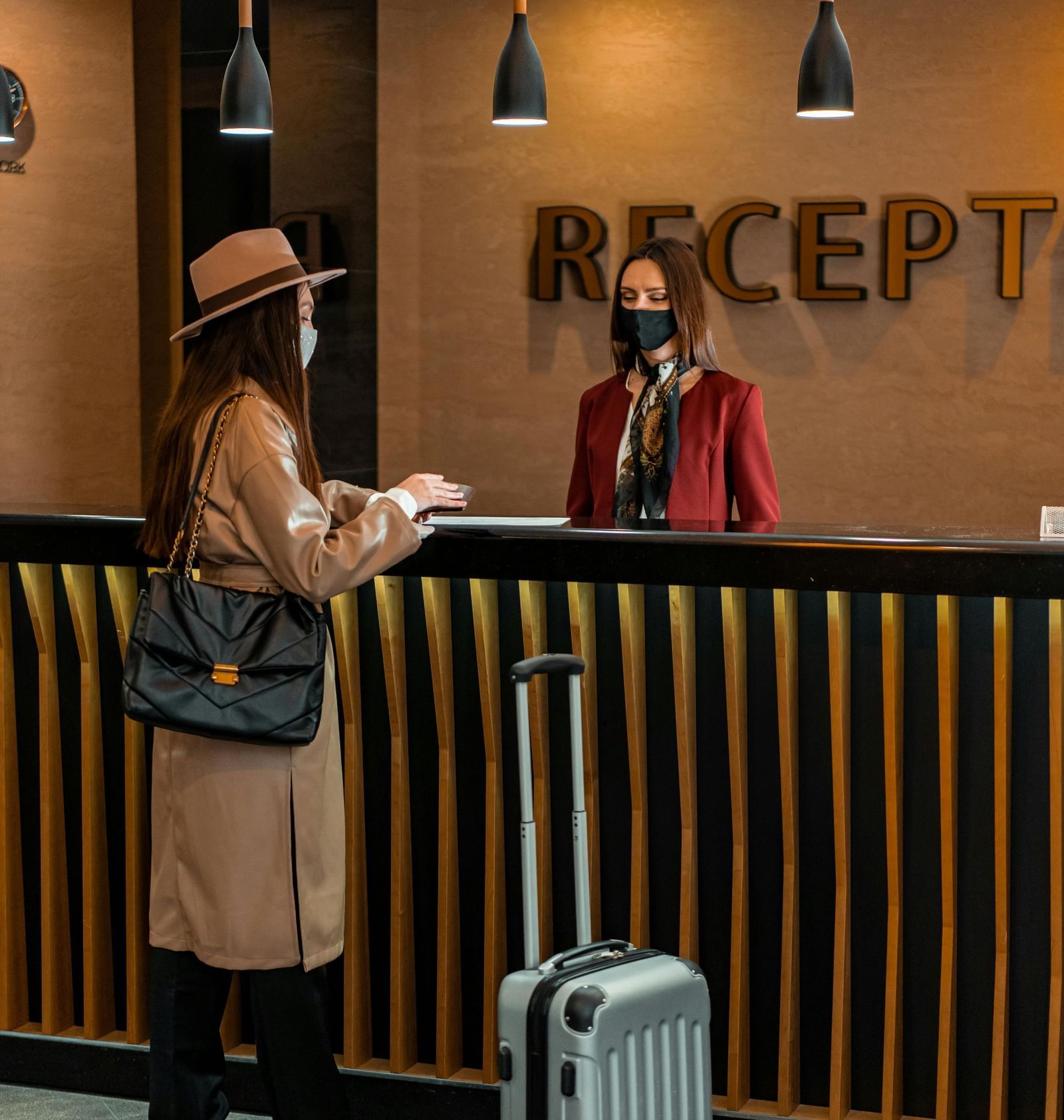 A receptionist standing behind the desk greeting the guest carrying a luggage trolley in front of her.