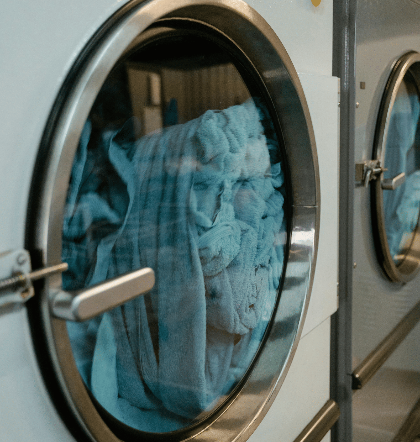 A row of industrial washing machines, with pale blue laundry visible through the stainless steel porthole doors.