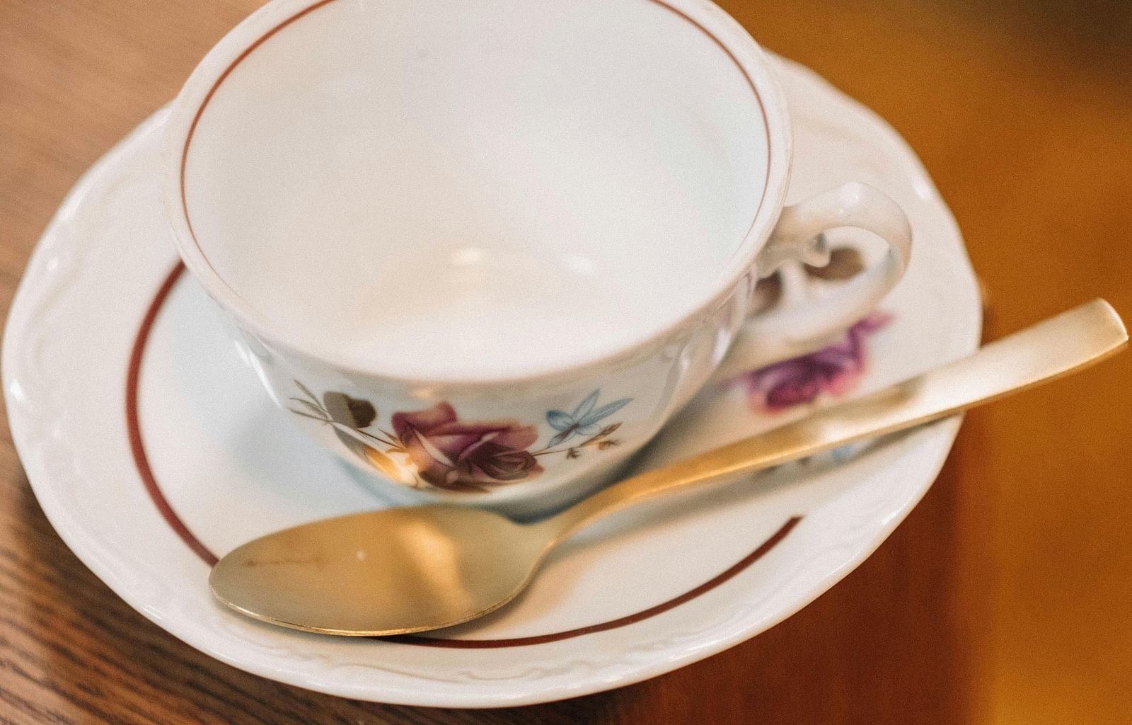 A white teacup with floral designs sits on a matching saucer with a red rim, accompanied by a golden spoon, all placed on a wooden surface.