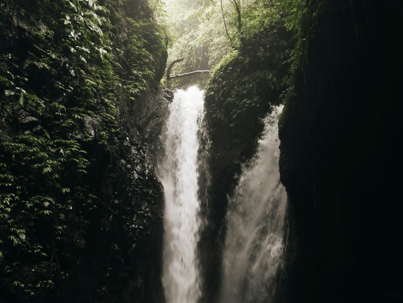 A twin waterfall pours into a dark, rocky cavern from a bright, leaf-framed opening at the top, creating a white pool.