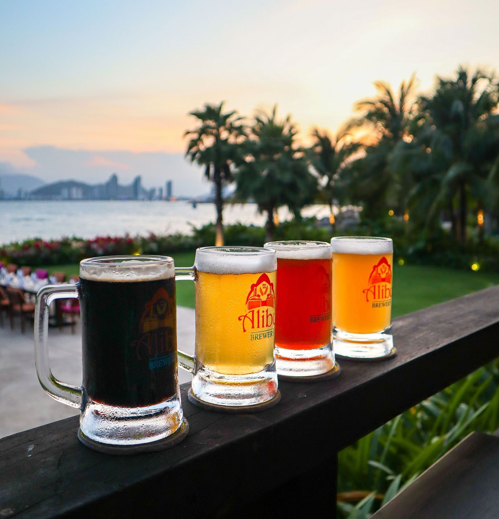 An array of drinks placed on a wooden railing near a beach - Alibu Resort Nha Trang