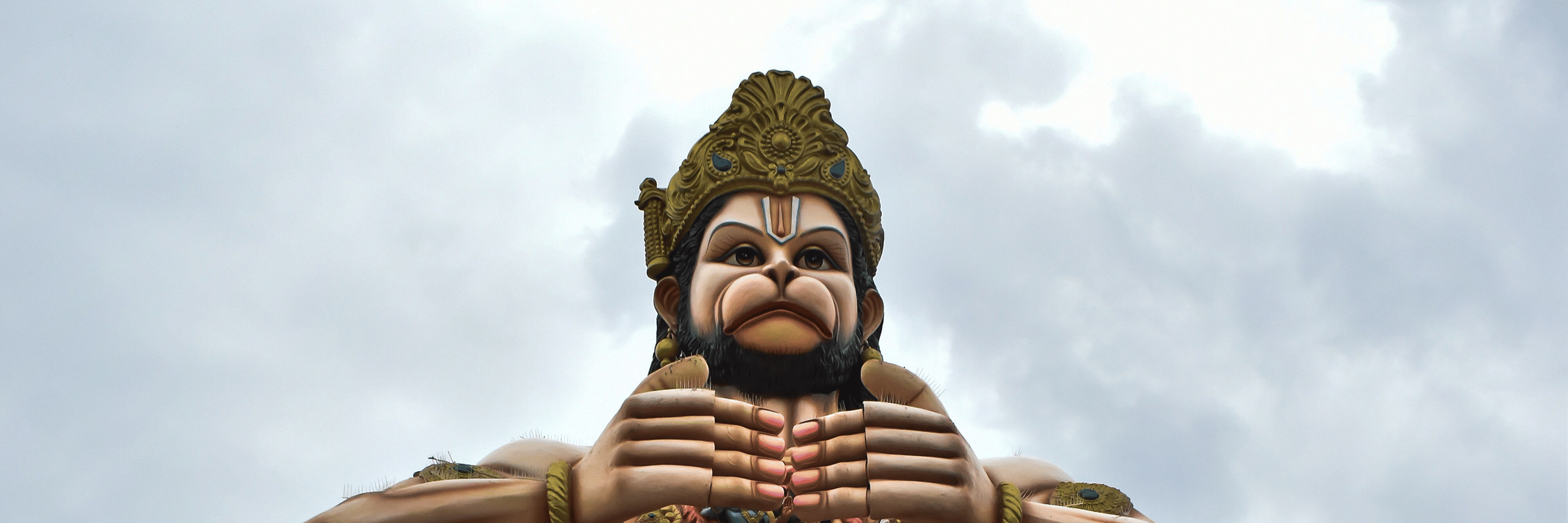 A large standing statue of a Hindu figure with a moustache and traditional attire, holding a mace against a cloudy sky.