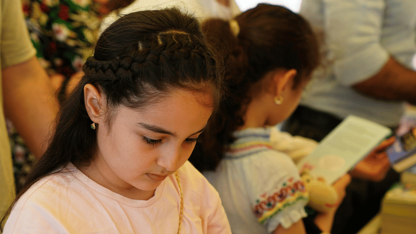 A young girl is focused on reading a book at a busy book fair or stall.