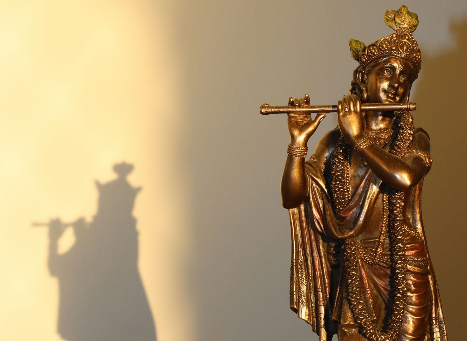 A golden statue of Lord Krishna playing the flute, casting a long shadow on a light wall.