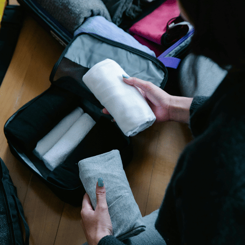 Hands packing neatly folded clothes into a suitcase on a wooden floor.