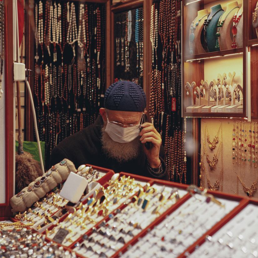 A view from outside of a shop keeper inside a jewellery store, wearing a mask and speaking on the phone, with many types of jewelleries kept to be sold.