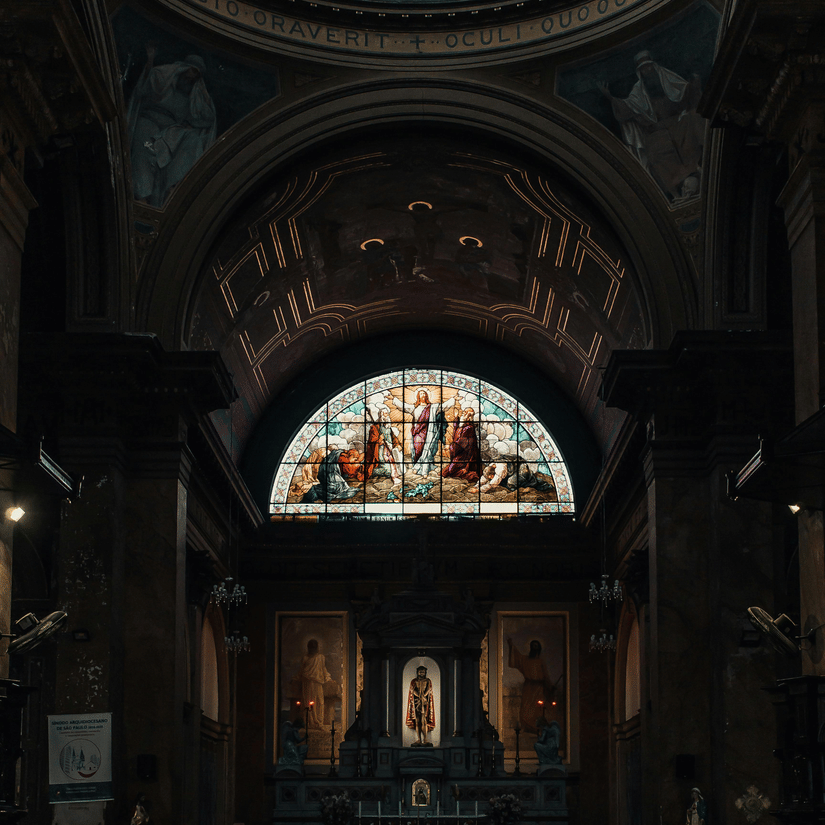 The dark interior of a cathedral shows a lit altar beneath a colourful semi-circular stained-glass window and dome.