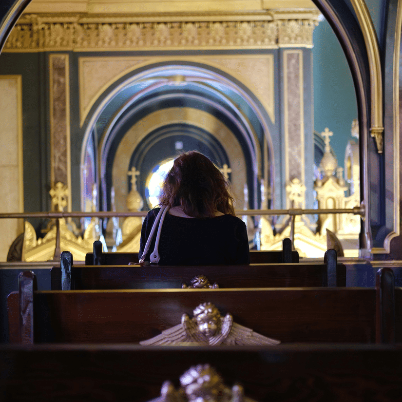 A woman sits in a dark wooden pew with angel carvings, looking toward the blue arched aisles of a grand cathedral.