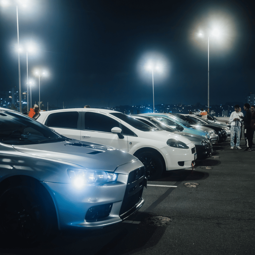 Several cars are parked closely together at night beneath the bright, harsh glare of tall street lamps.