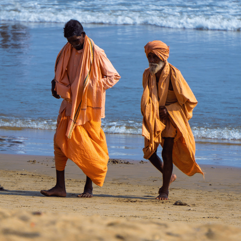 Two men in saffron dress walking on the beach with the waves of the waterbody clashing against the beach.