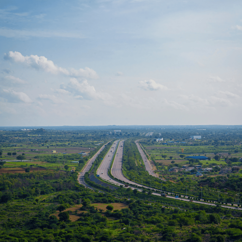 An aerial view of a wide, multi-lane highway stretching through a lush, green landscape under a bright, partly cloudy sky.