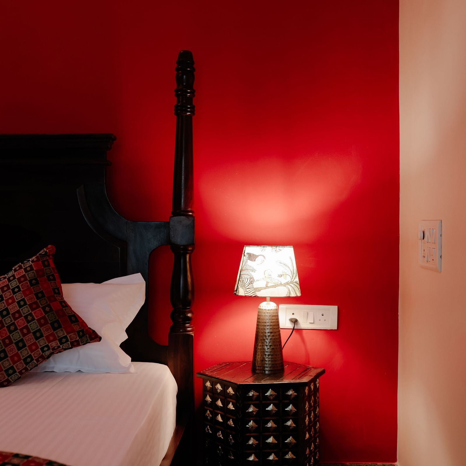 A glimpse of a bed with patterned linens, a bedside table with a lamp, and a bold red wall | Adhyaya Heritage Reverbed