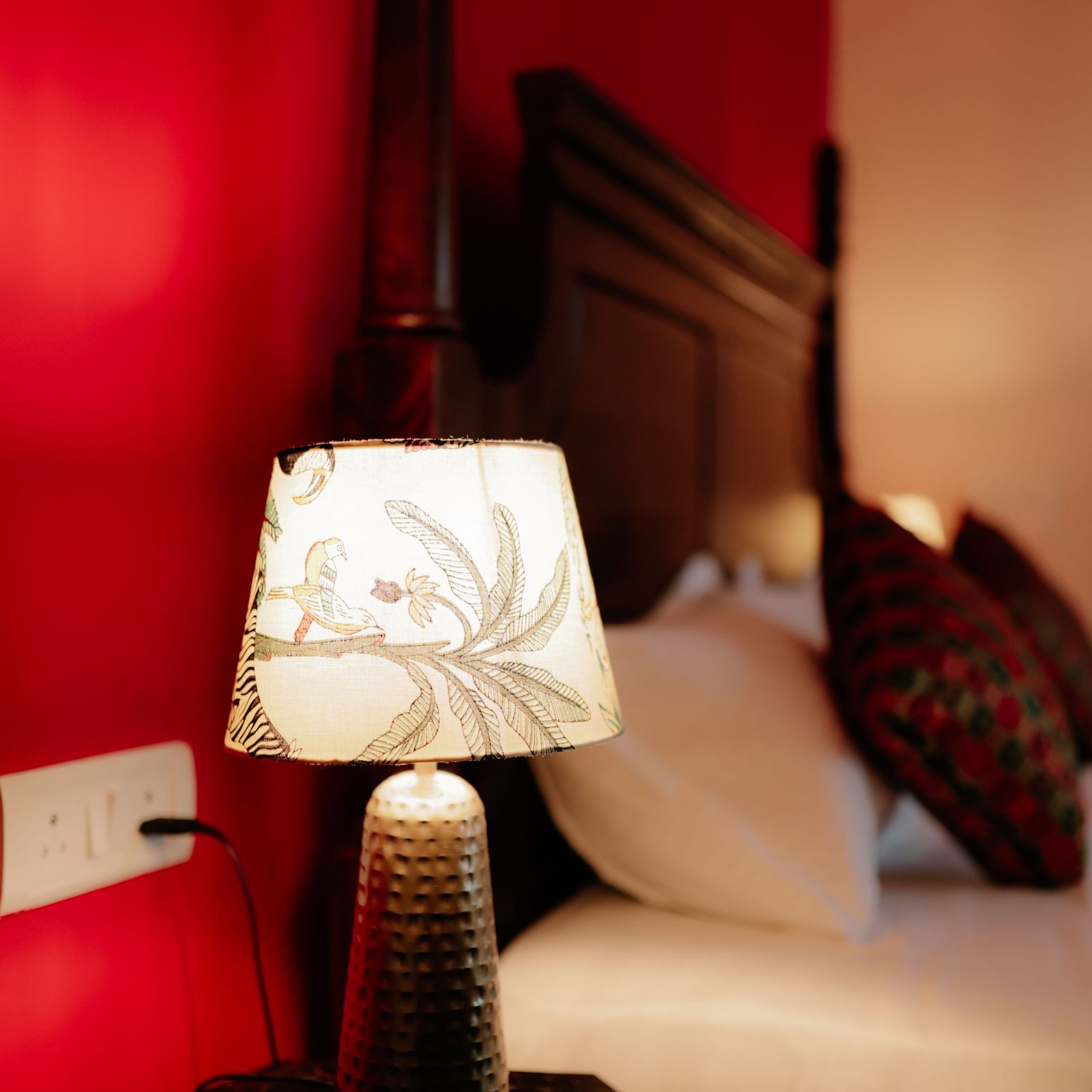 A bedside lamp illuminating a portion of a bed with white linens and a patterned throw, against a bold red wall | Adhyaya Heritage Reverbed
