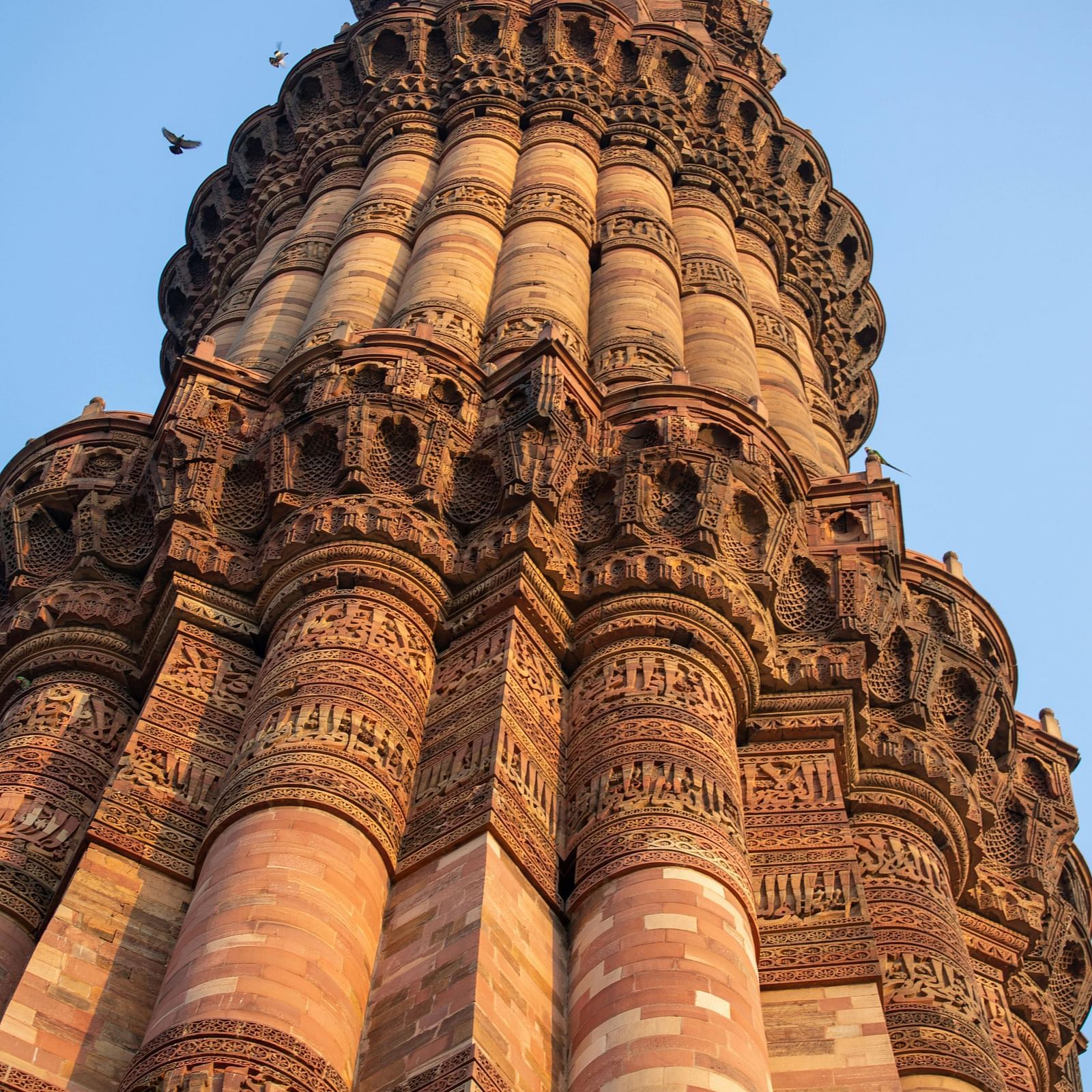 A low-angle view of the Qutub Minar, a tall, intricately carved, reddish-brown minaret, stands against a clear blue sky.