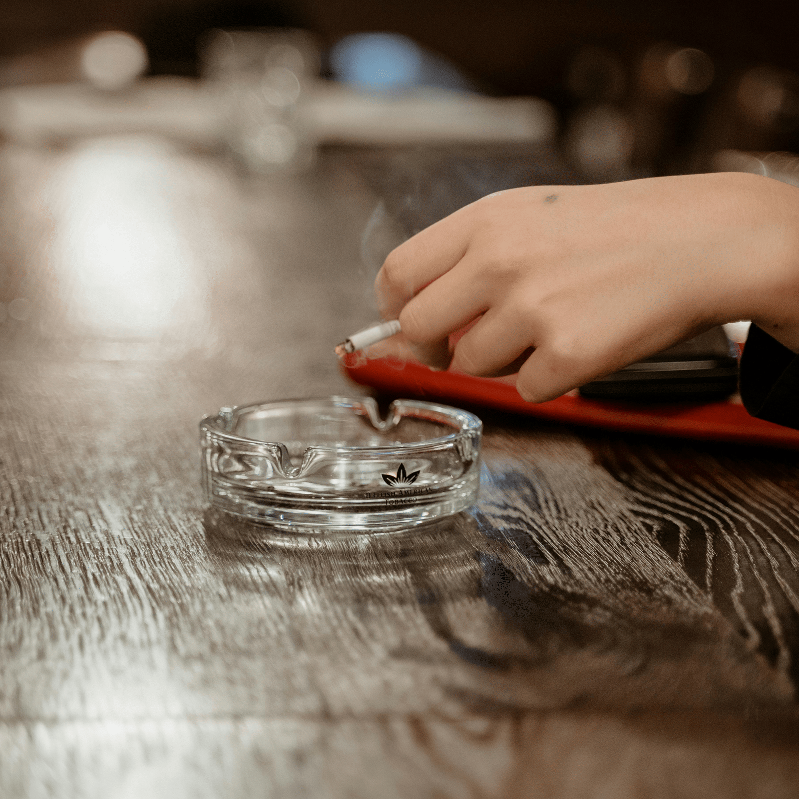 A person holding a cigarette over a crystal ash tray kept on a counter. 