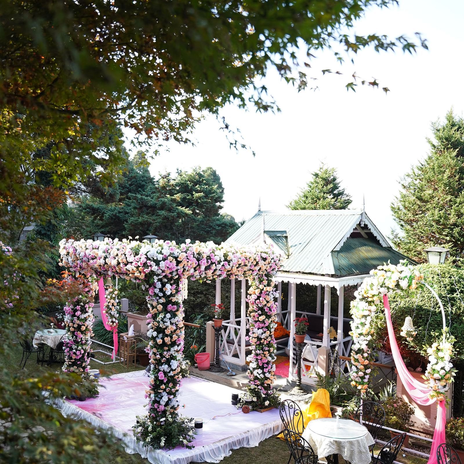 The outdoor wedding ceremony setup featuring a flower adorned pavilion and small seating tables surrounded by tall trees at the Cedar Inn in Darjeeling.