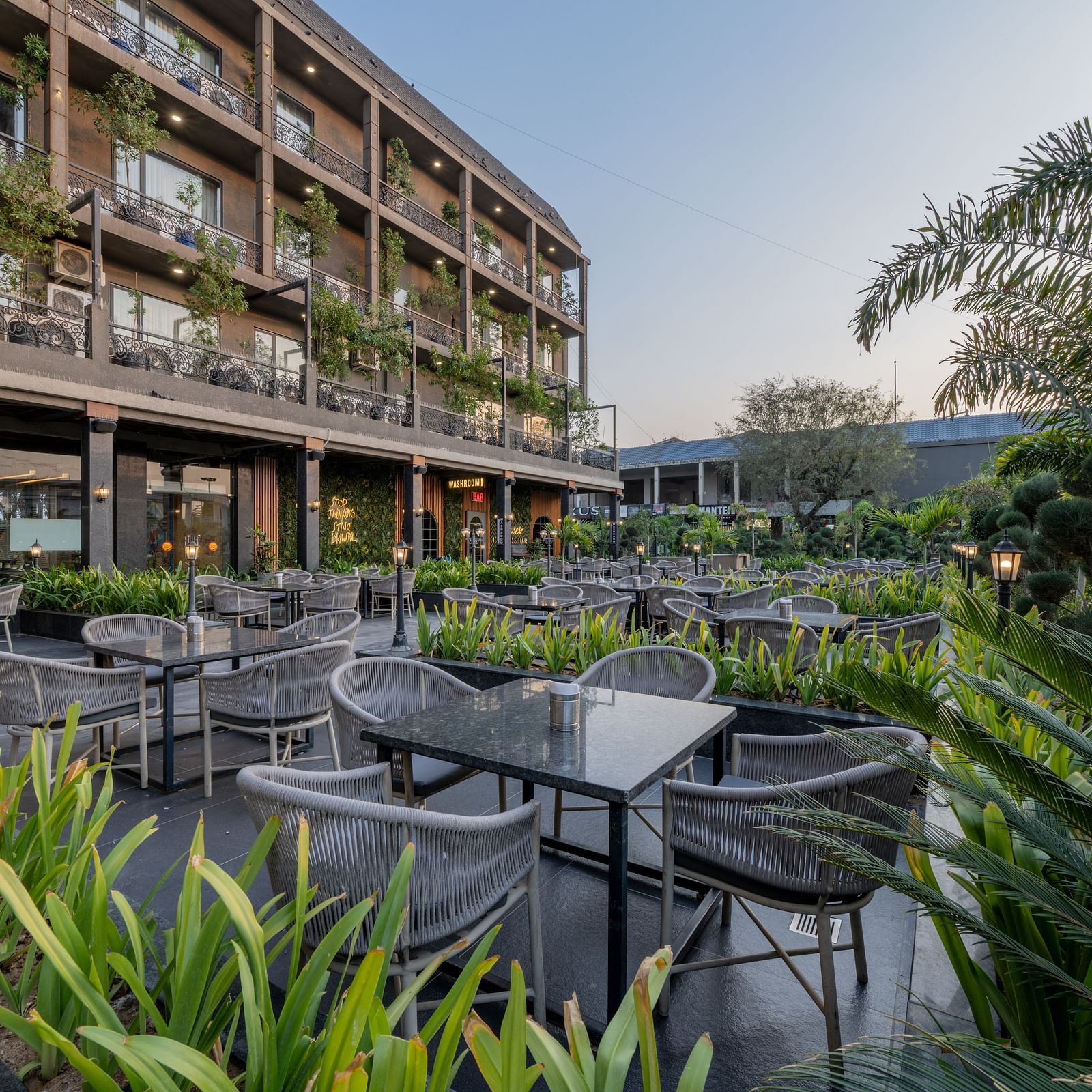 Outdoor seating area at Hotel Highway King, Bagru, with tables and chairs surrounded by plants and hotel balconies.