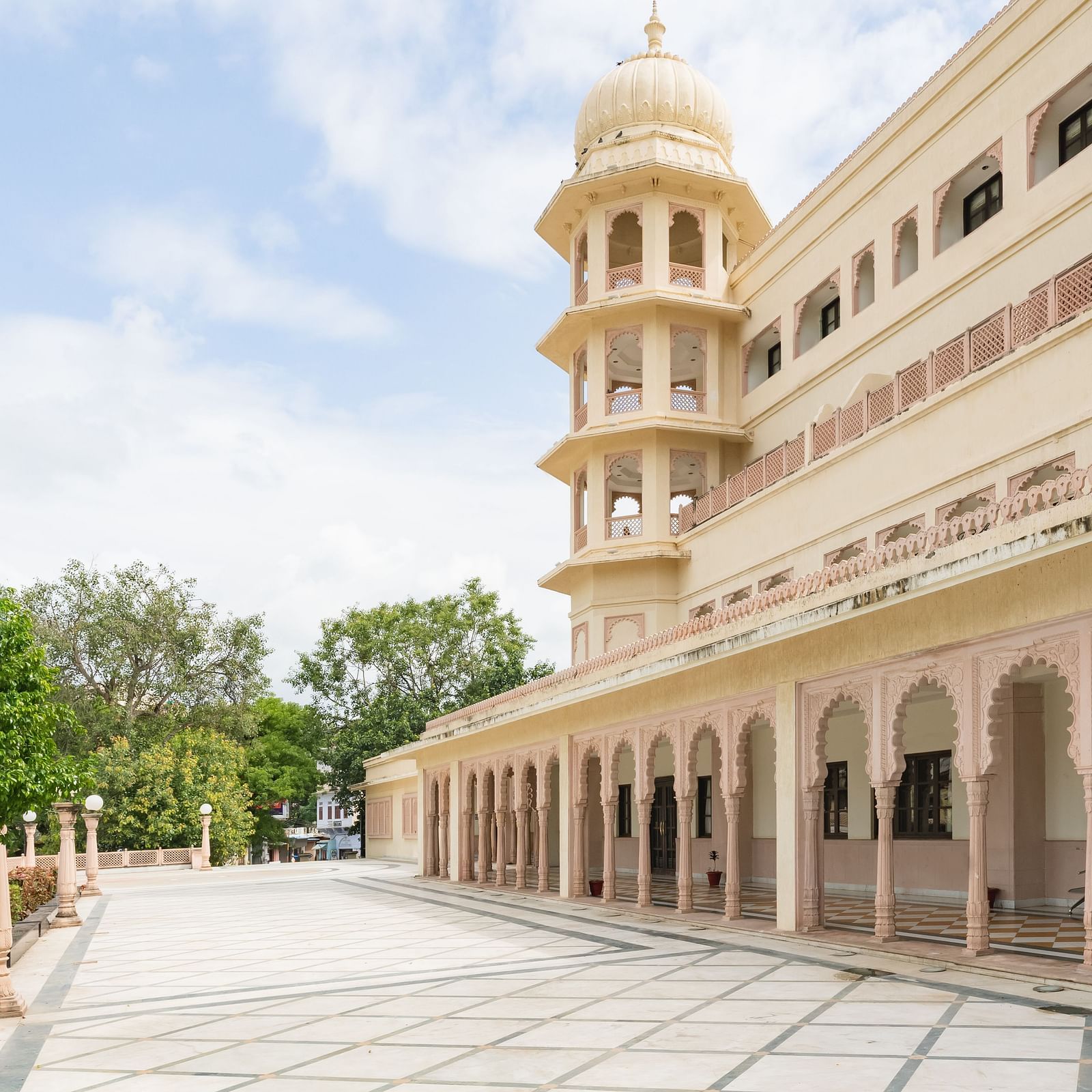 The facade of Shri Vallabh Vislas Lords Plaza Nathdwara featuring patterned white tiles in the foreground and lush green vegetation under blue sky at the backdrop.