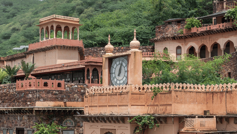 Historic stone buildings topped with a clock tower sit against a lush green hill in traditional architecture.