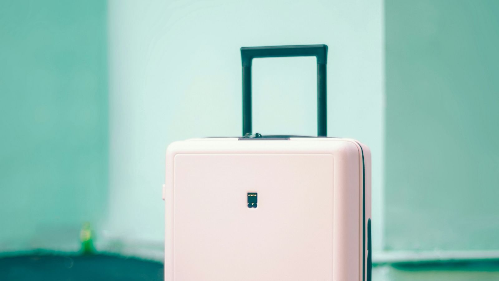 A pastel pink suitcase with a retractable handle, standing upright on a dark green floor with white markings, against a teal wall.