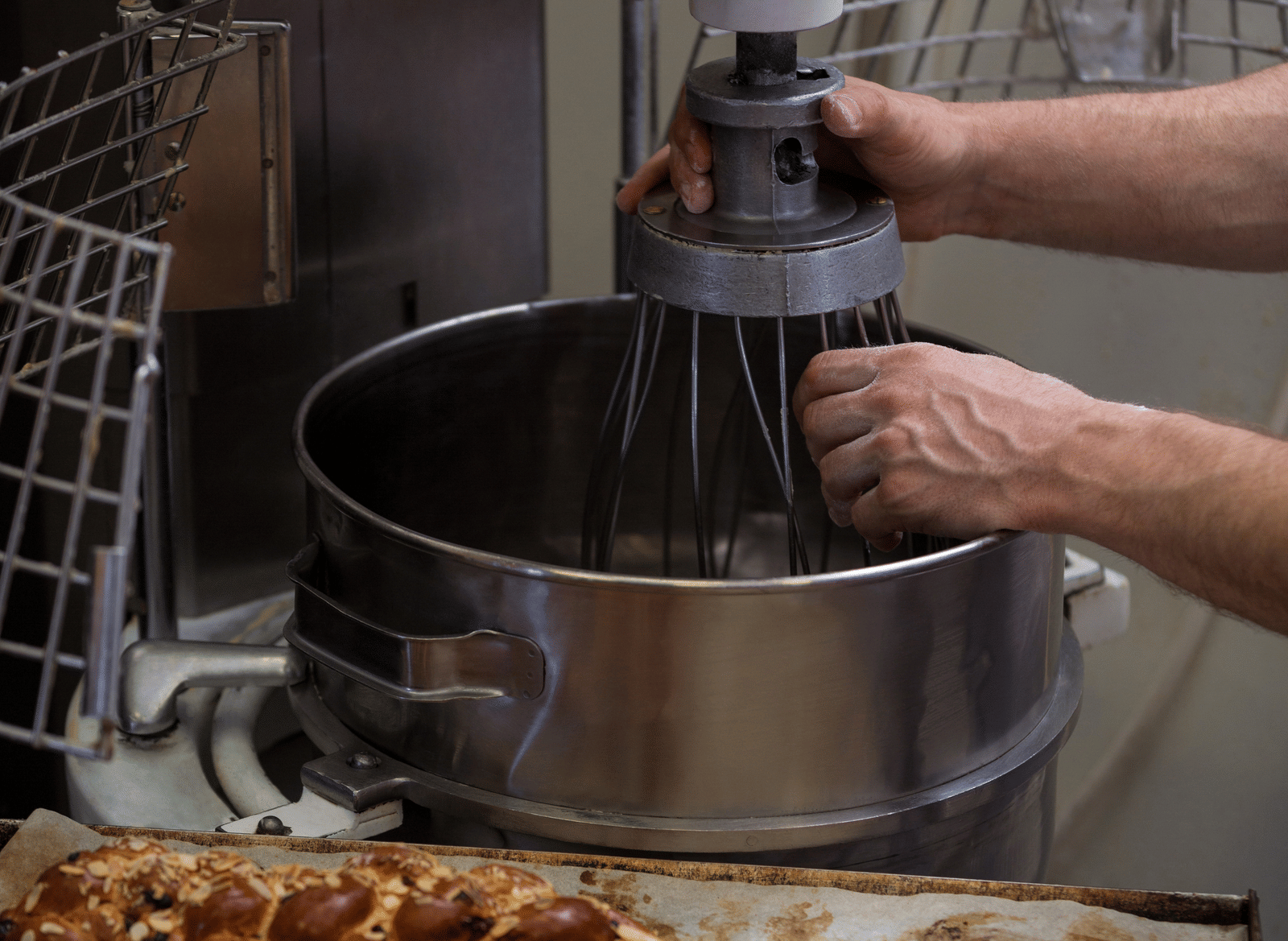 A person adjusts the whisk attachment on a large industrial mixer in a bakery, with trays of freshly baked braided bread topped with nuts in the foreground.