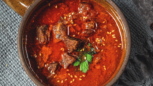 a bowl of mutton curry with garnishing kept on a table with a fork and knife next to it