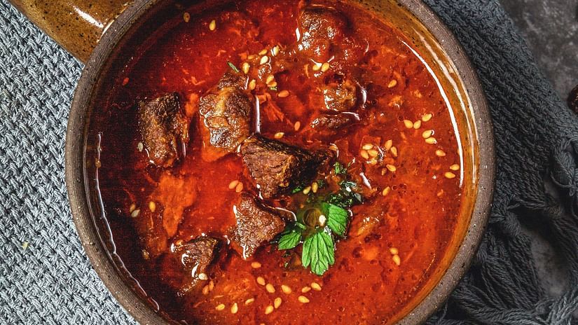 a bowl of mutton curry with garnishing kept on a table with a fork and knife next to it