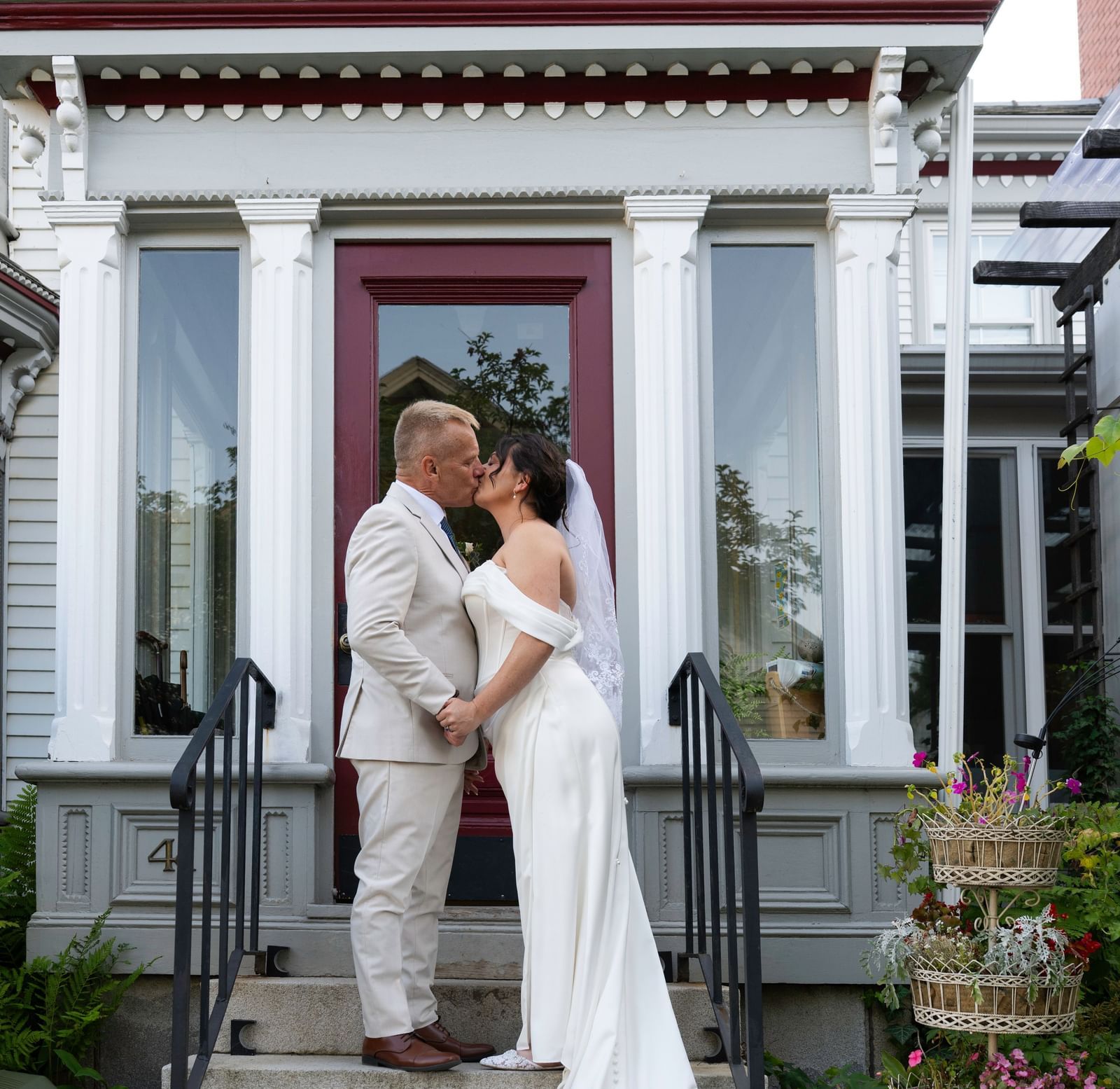 A bride and groom kiss while standing on the steps in front of a doorway at Hartstone Inn, holding hands with flowers and plants visible beside them.