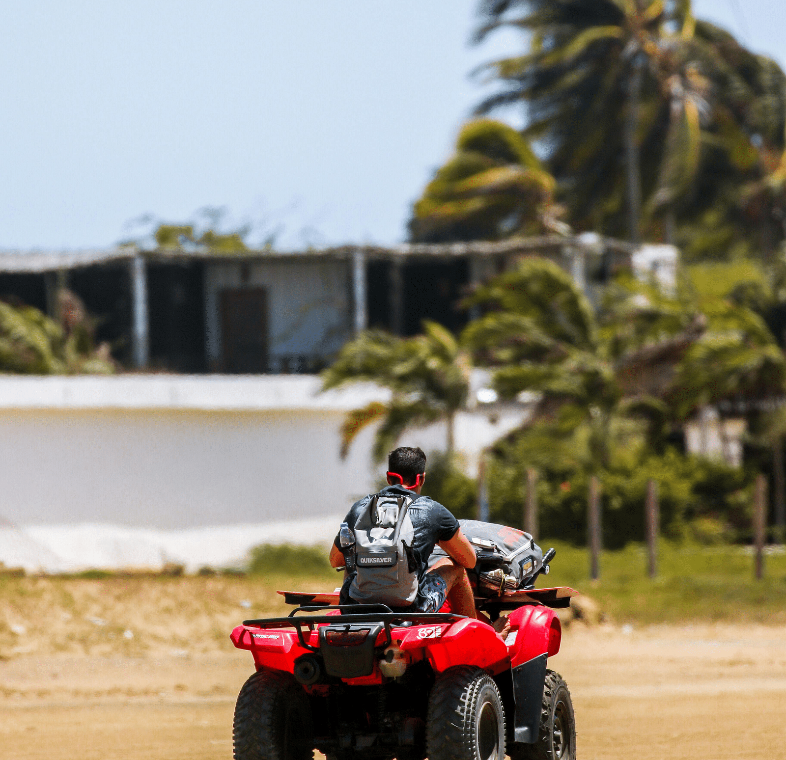 A person riding a red quad bike on a sandy area with sparse vegetation and a building in the background, under a clear sky.