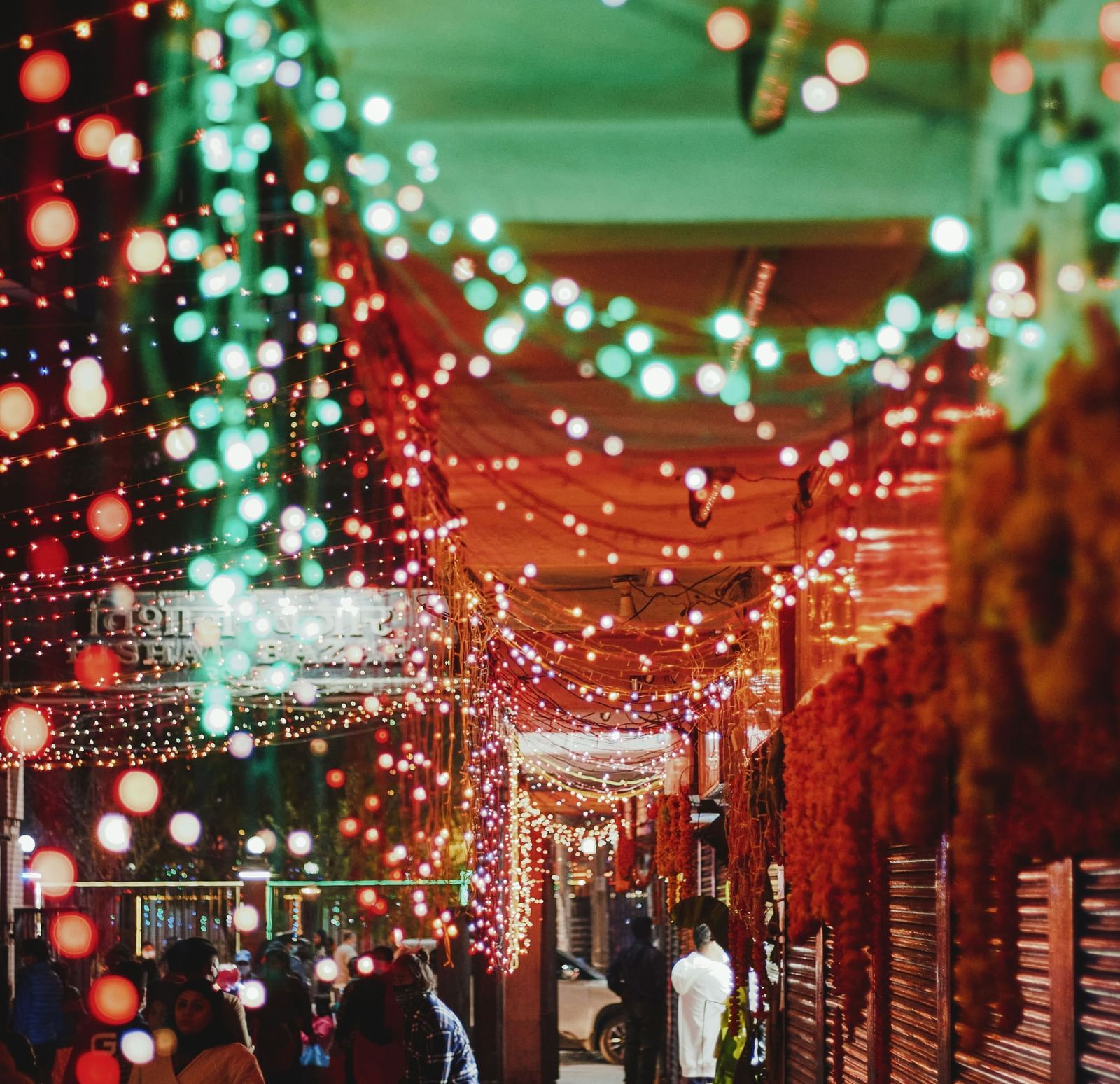 A vertical shot of a bustling, narrow outdoor alley decorated with a dense canopy of cascading fairy lights in warm red and bright aqua-green, with people walking beneath the glowing lights.