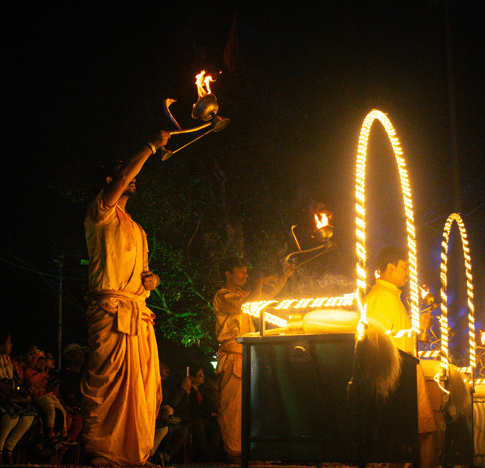 three priests doing aarti during late evening as the devotees are sitting in the background
