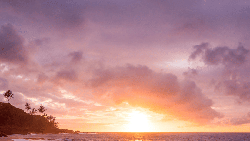 The shore of a beach during sunset, with waves reflecting the warm colours of the sky.
