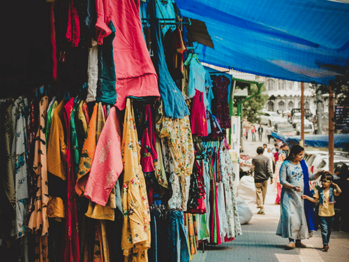 Colourful clothing displayed on racks under a blue canopy at a sunny outdoor street market with pedestrians.
