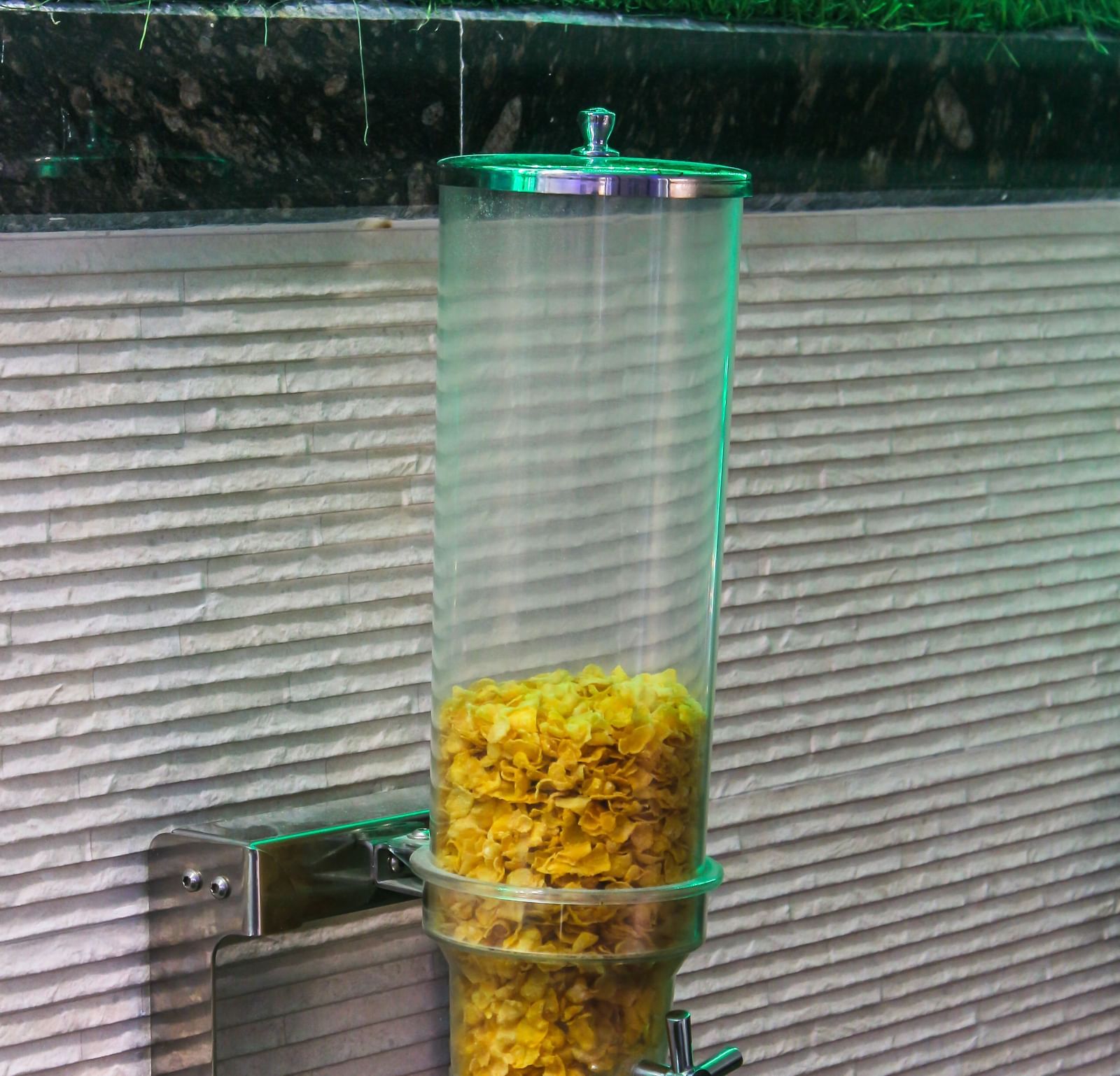 A clear plastic dispenser filled with cereal, mounted on a textured wall with green lighting above. - Hotel Diamond Plaza, Chandigarh.