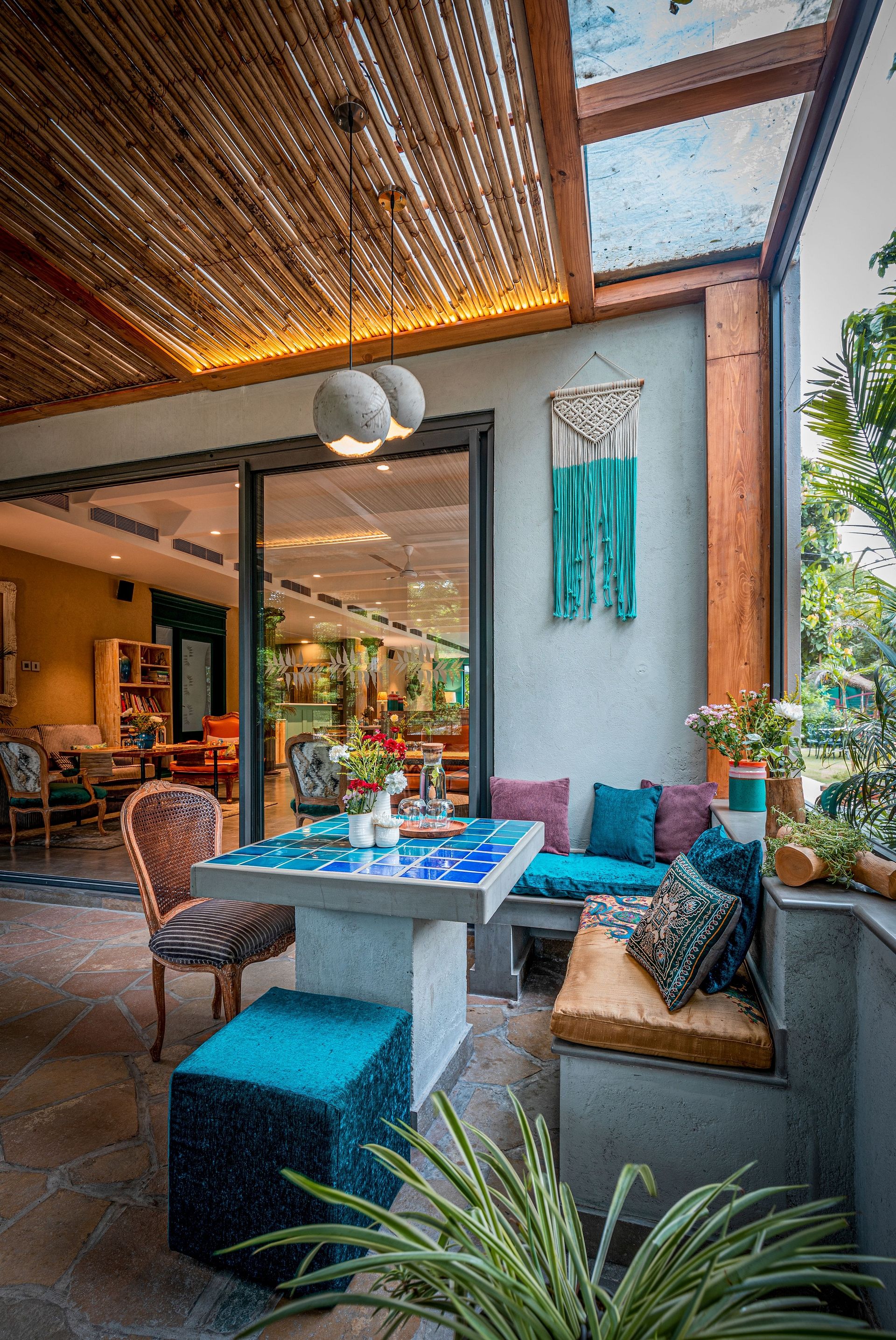 Under The Neem cafe area with a blue and wooden theme, featuring a dining table, bench cushions, plants, and a glass ceiling letting in natural light at Karma Chalets.