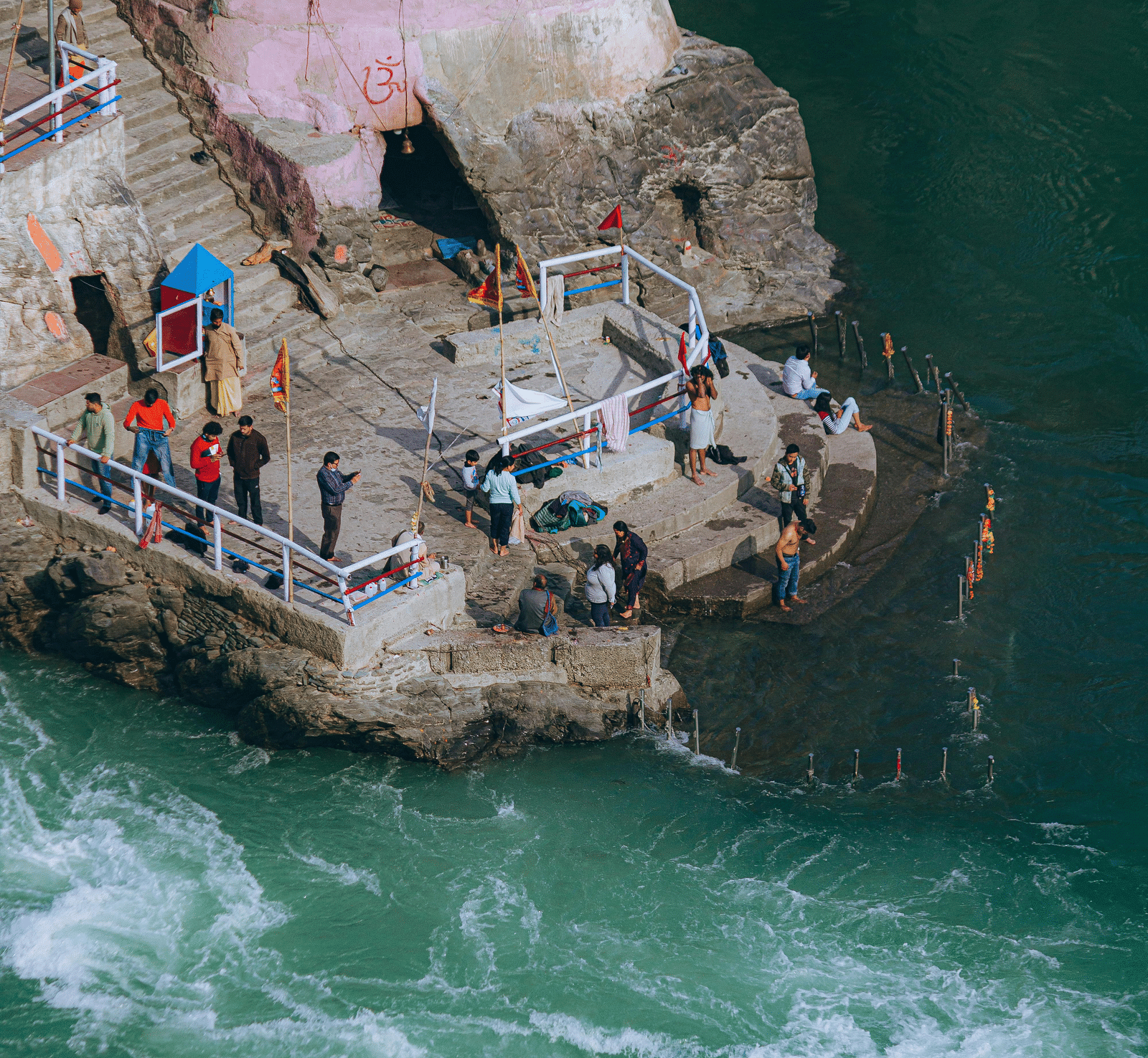 A high-angle view of a confluence, with people gathered on a stone platform by swirling, green waters, adjacent to a building with a pink facade.