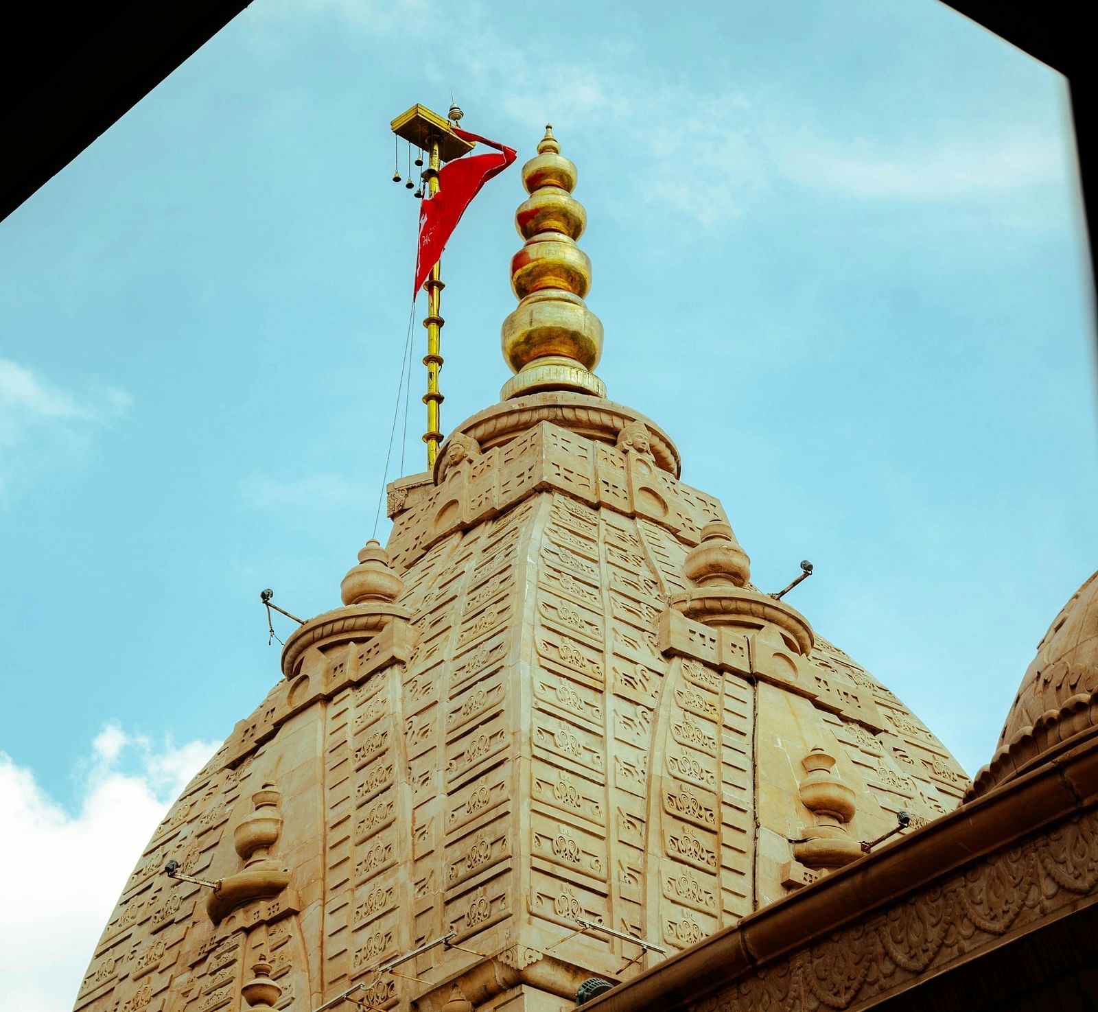 A stone temple spire with golden finials and a red flag, viewed through a triangular frame against a blue sky.