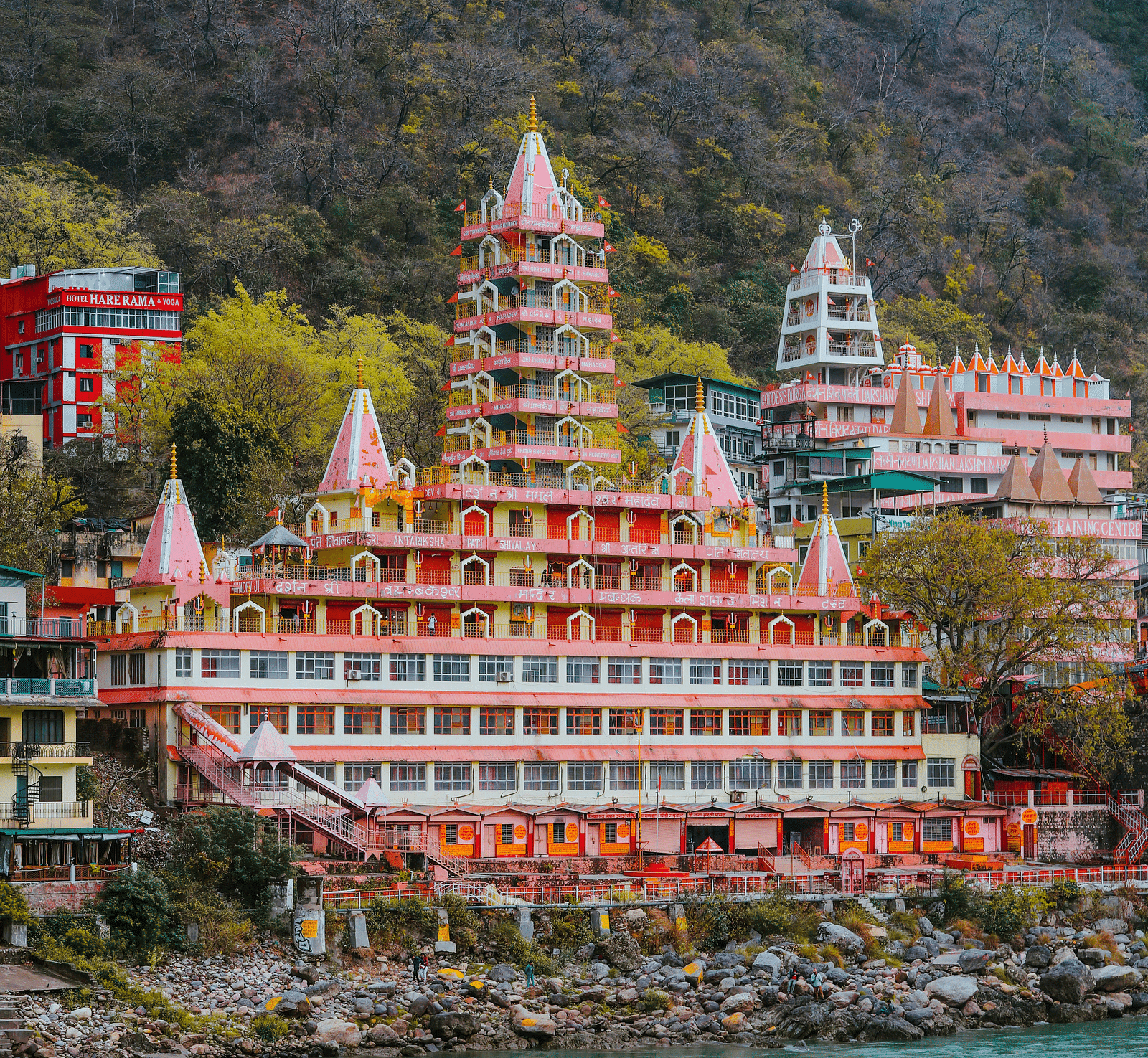 A multi-tiered temple with vibrant red and orange tones stands beside a flowing river, encircled by trees.