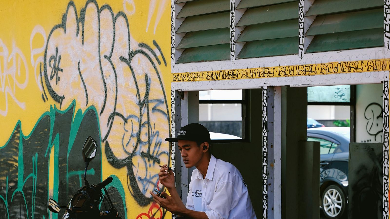 A person sitting on a window sill next to a graffiti with his motorbike parked in front of him.