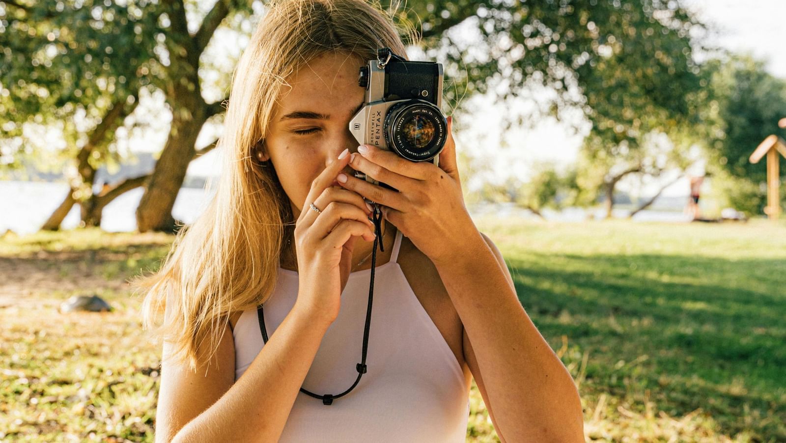A woman with a camera, clicking pictures with sunlight, grass and trees behind
