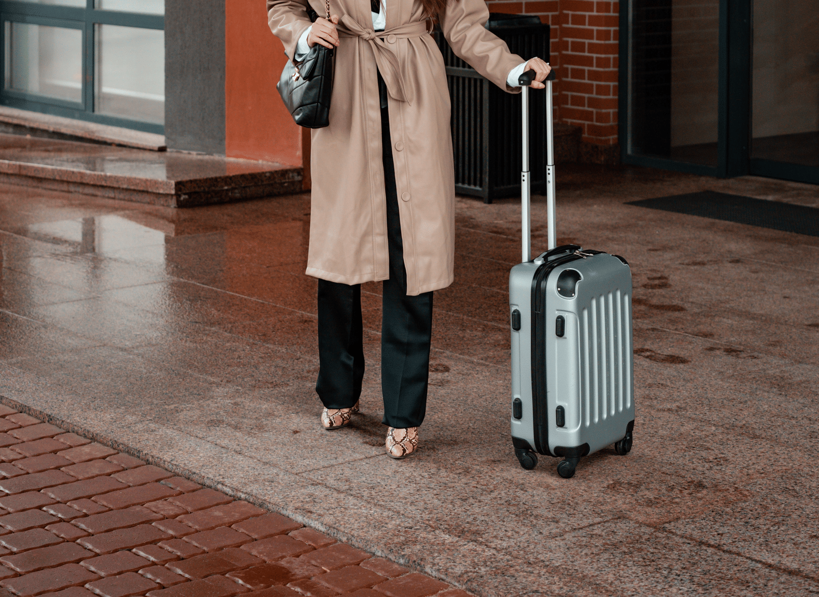 A person wearing a hat, face mask and coat walks outdoors along a pavement next to a building while pulling a wheeled suitcase.