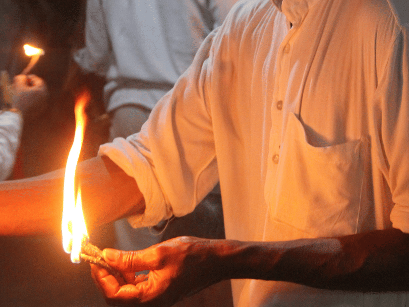 A man in a white shirt holding a small flickering flame during a religious ceremony with pink drapes in the background.