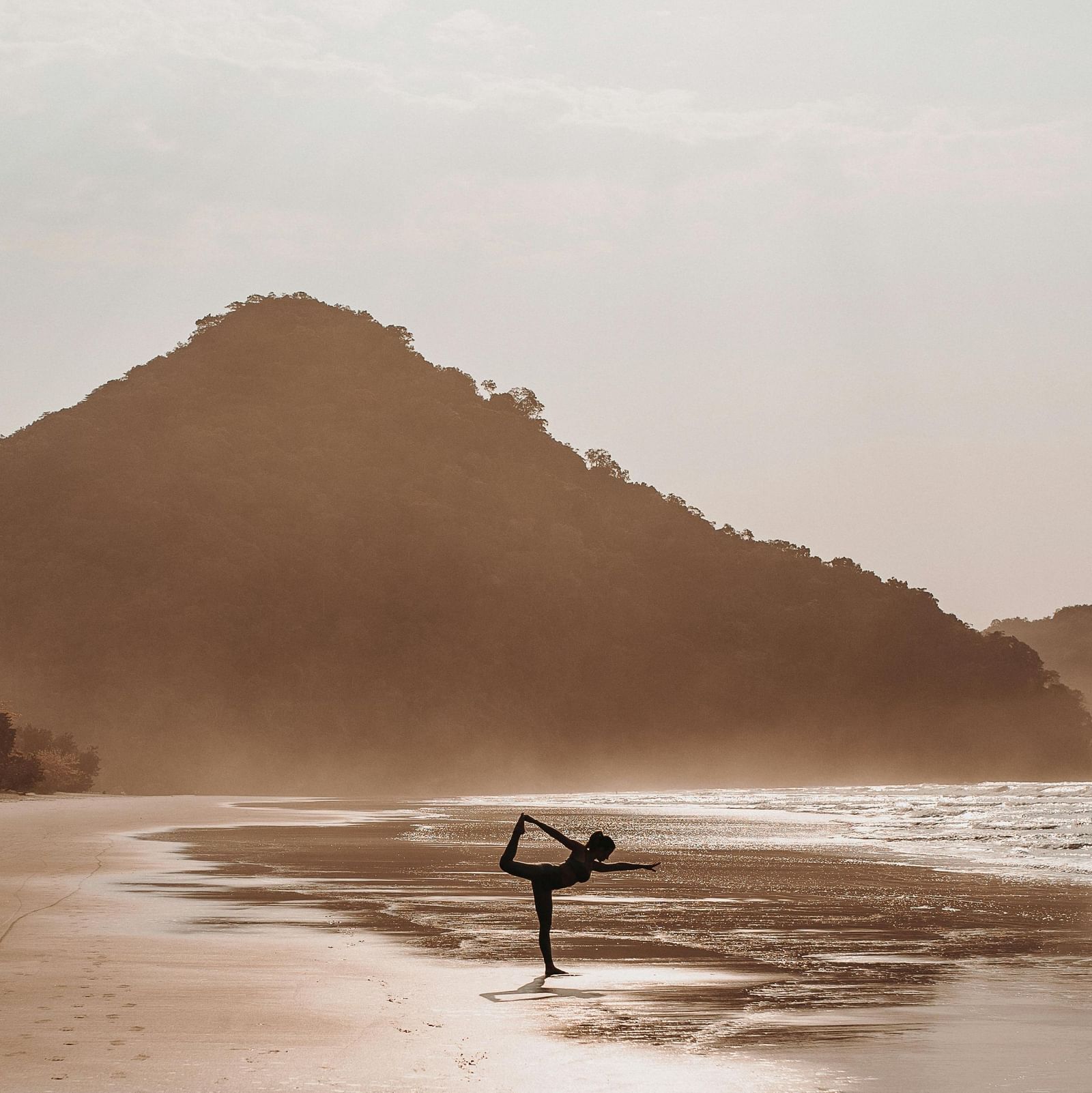 A woman doing yoga on a beach facing towards the sea along with a hill like structure in the background.