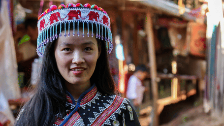Young woman in embroidered black tribal dress and beaded red elephant-motif headdress standing in a sunlit village market lane.