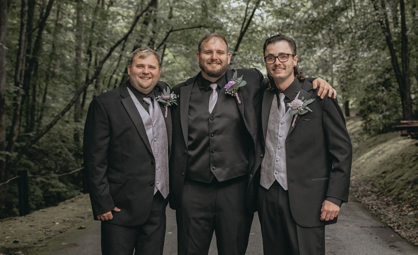 Groom standing with two men on either side embracing each other on a road covered with trees.