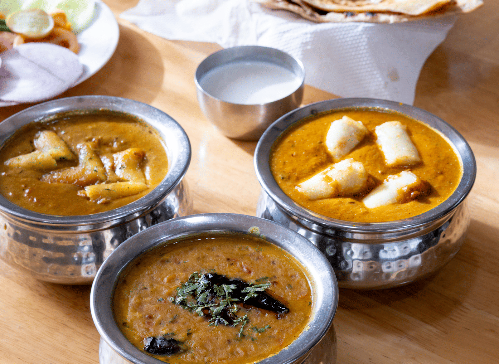 Traditional Indian food: metal bowls with curry and dal, with flatbread (roti) and fresh salad on a wooden surface at Pravasa Stay, Vadodara.