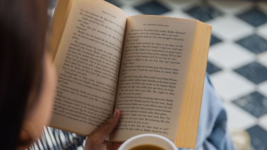 Person seen from behind reading a book and sipping coffee in a sunlit corner with checked flooring, reflecting the cozy leisure options available at The Manor Sports & Wellness Hotel in Shimla