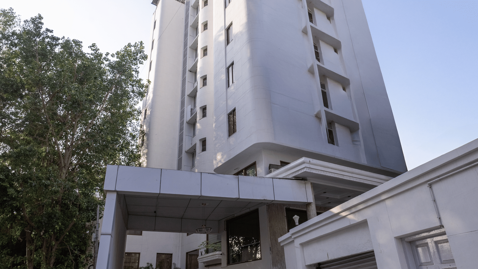 Exterior view of the tall, white hotel building standing against a clear sky, with a driveway leading to the entrance at Pravasa Stay, Vadodara.