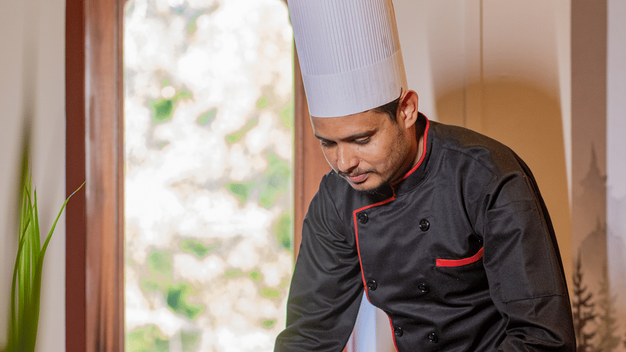 A chef in a black uniform and white hat arranging a dish on a table inside Estherea Bagh, Ranthambore.